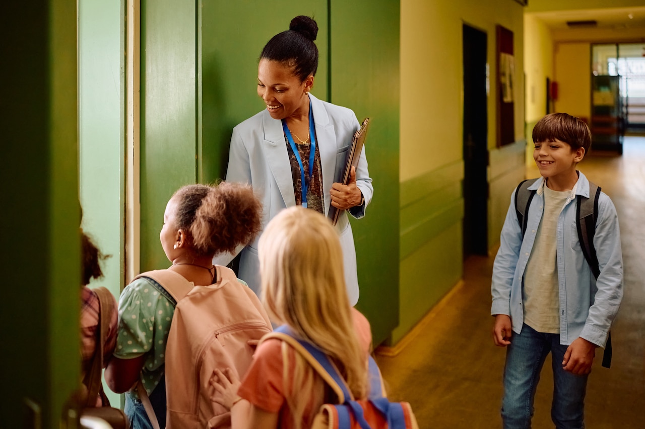 Happy black teacher greeting elementary students in front of the classroom.
